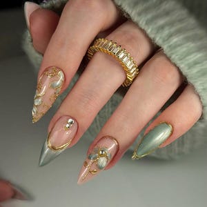 Op de afbeelding: Close-up van een hand met lange, puntige nagels met ingewikkelde nail art. De nagels tonen een verscheidenheid aan ontwerpen, waaronder gouden accenten, bloemversieringen en een gouden ring met rechthoekige stenen. De nagels zijn geschilderd in roze, zilver en groen.