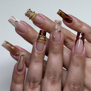 May include: A set of long, square-shaped acrylic nails. The nails feature a variety of designs, including a French tip, gold embellishments, and a tortoiseshell pattern. Some nails have gold rings and small gold studs.