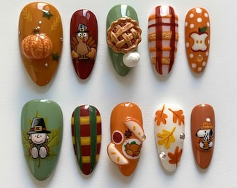 May include: A set of ten almond-shaped artificial nails with a fall theme. Designs include a 3D pumpkin, turkey, pie, plaid, apple, Snoopy, and fall leaves. Colors include orange, green, red, and brown.