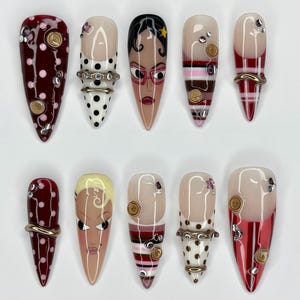 May include: A set of ten stiletto-shaped press-on nails featuring diverse designs including maroon polka dots, black and white patterns, striped motifs, and two nails painted with stylized character faces, all accented with small buttons and rhinestones.