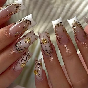 May include: Long, square-shaped acrylic nails with a white tip and a gold filigree design. Some nails feature gold flower embellishments. The nails have a clear base with a gradient effect.