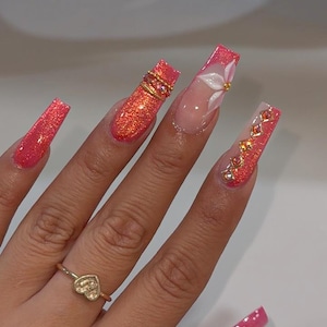 Pode incluir: Unhas longas de formato quadrado com um acabamento de glitter coral vibrante. O design inclui detalhes florais brancos em relevo, anéis metálicos dourados e pequenos cristais cor de âmbar incrustados. Um anel fino de ouro com um coração é usado.