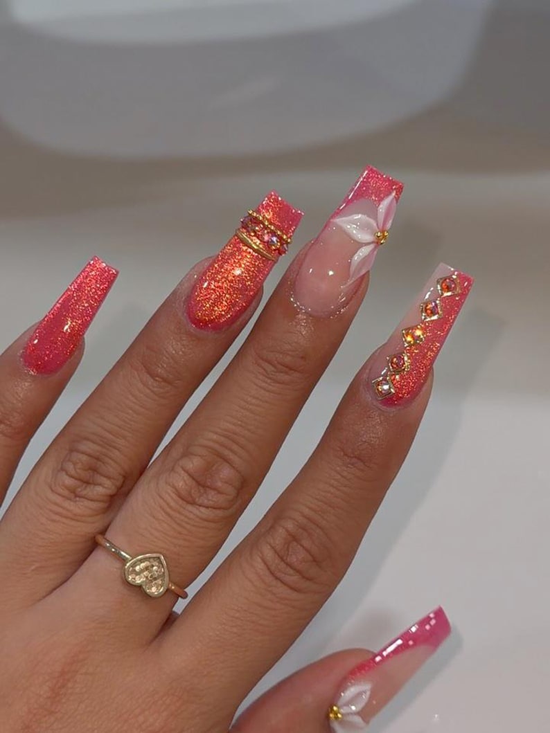 May include: Long square-shaped nails featuring a vibrant coral glitter finish. The design includes three-dimensional white floral accents, gold-toned metal rings, and small embedded amber-colored crystals. A thin gold ring with a heart charm is worn.