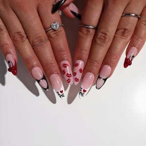 May include: A close-up of manicured nails with various designs. Some nails feature a cherry design, others have red lips, and some have the text "I ❤️ ME". The nails are long and almond-shaped, with a pink and white color scheme.