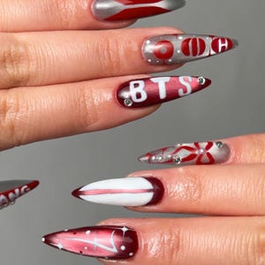 May include: A set of long, pointed fingernails with various designs. The nails feature a combination of red, silver, and white colors, with some adorned with text like "BTS" and "ARMY". Some nails have decorative rhinestones.