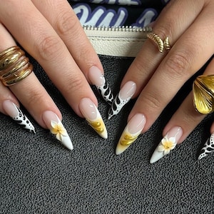 May include: Close-up of manicured nails with a variety of designs. Some nails feature a white base with black and white patterns, while others have gold accents and 3D flower details. The nails are long and pointed.
