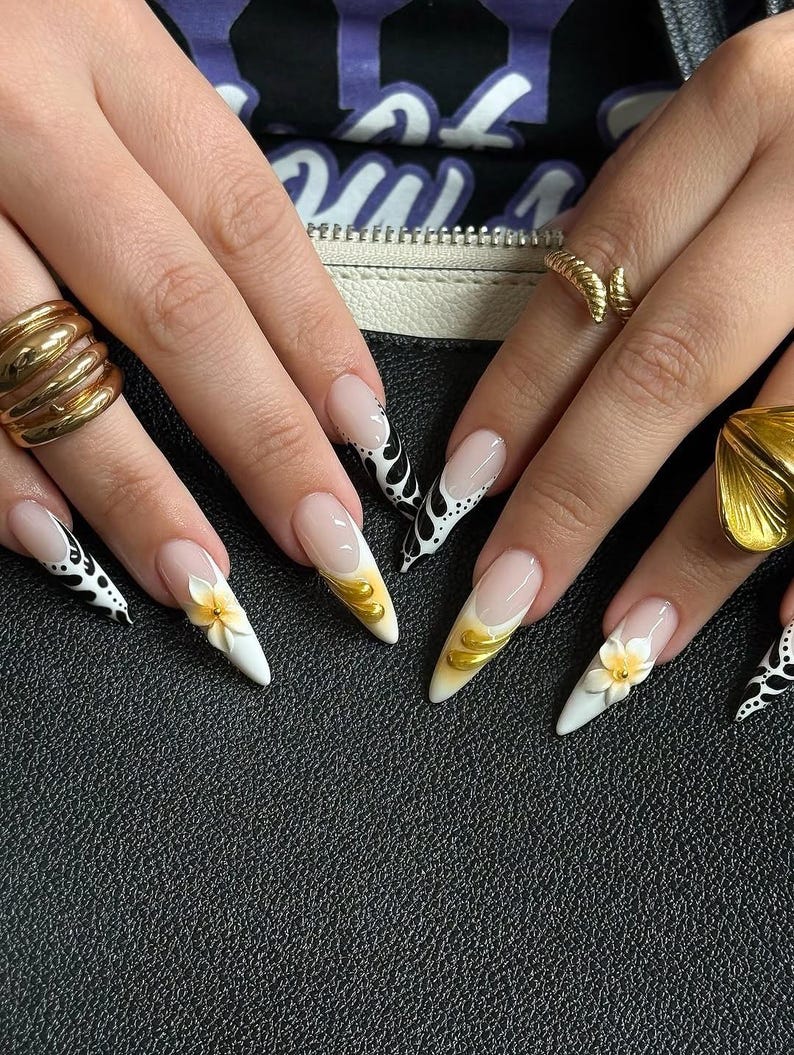 May include: Close-up of manicured nails with a variety of designs. Some nails feature a white base with black and white patterns, while others have gold accents and 3D flower details. The nails are long and pointed.