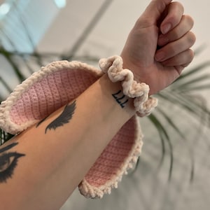 May include: A light pink crochet bunny ear headband with a cream-coloured trim. A cream-coloured scrunchie is worn on the wrist. The arm has a tattoo of the number 777 and an eye.