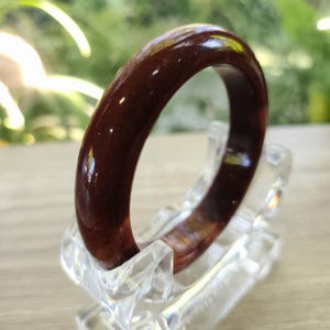 May include: A polished, dark brown bangle. The circular bracelet is displayed on a clear, angled stand. The smooth surface and glossy finish of the bracelet are highlighted against a blurred green background.
