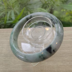 Könnte beinhalten: Ein Jade-Armreif mit Grün- und Schwarztönen. Der Armreif ist rund und glatt, mit polierter Oberfläche. Der Jade ist durchscheinend und lässt Licht durch. Der Armreif wird auf einem transparenten Ständer präsentiert.