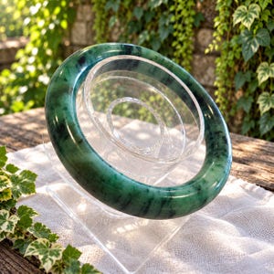 May include: A smooth, polished bangle bracelet featuring a deep green color with dark, mottled inclusions throughout the stone. The circular bangle has a thick, rounded profile and is displayed on a clear, tiered acrylic stand.
