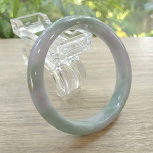 May include: A jade bangle bracelet with a smooth, polished surface. The bracelet features a blend of pale green and lavender hues. The jade is displayed on a clear, decorative stand, showcasing its natural beauty. The bangle's diameter is approximately 6.35 cm.