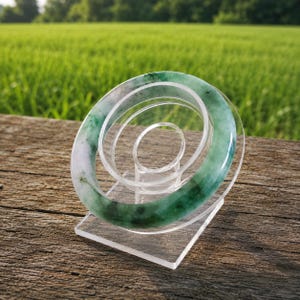 Puede incluir: Un brazalete circular de jade con una superficie lisa y pulida de aspecto translúcido. El brazalete muestra un patrón variado de verde bosque profundo e inclusiones minerales blancas a lo largo de toda su estructura de anillo sólido.