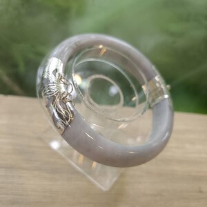 以下が含まれることがあります： 滑らかな淡いラベンダーグレーの石製バングルブレスレット。片側には、複雑なフィリグリースタイルの彫刻が施された光沢のあるシルバー調の金属製オーバーレイがあしらわれています。円形のバンドは丸みを帯びたプロファイルで、光沢のある反射仕上げが特徴です。