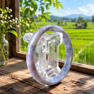 May include: A smooth, circular bangle bracelet crafted from polished lavender and grey stone with natural marbling patterns. The bangle is displayed upright on a clear acrylic stand, showcasing its translucent quality and rounded, polished surface.