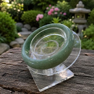 Könnte beinhalten: Ein jadegrünes Armreif-Armband, das auf einem transparenten Acrylständer ausgestellt ist. Das Armband hat eine glatte, polierte Oberfläche. Der Ständer hat ein geometrisches Design mit einer kreisförmigen Oberseite und einer quadratischen Basis. Der Hintergrund zeigt Grün und eine Steinlaterne.