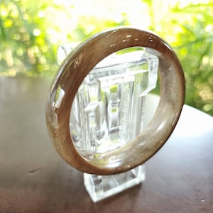 May include: A translucent brown bangle bracelet. The bracelet is displayed on a clear glass stand, with a blurred green background. The bangle has a smooth, polished surface and a circular shape. The color is a mix of light and dark brown.