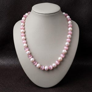 Op de afbeelding: Een ketting van afwisselend roze en witte kralen, tentoongesteld op een mannequin. De ketting heeft ronde, lichtroze kralen en grotere, crèmekleurige parels. Een delicaat sieraad.