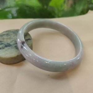 Op de afbeelding: Een gladde, gepolijste armband gemaakt van doorschijnende lichtlavendelkleurige en bleekgroene steen. De cirkelvormige band heeft een subtiele, donkerpaarse insluiting aan één kant en toont een natuurlijk, afgerond profiel met een glanzende afwerking.
