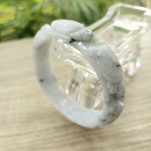 Puede incluir: Brazalete de jade blanco con diseños de tortugas y olas talladas. El brazalete tiene un acabado liso y pulido con sutiles inclusiones verdes y negras. Las tallas de tortugas son detalladas.