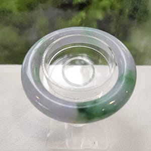 Pode incluir: Uma pulseira circular lisa feita de pedra translúcida, com uma superfície polida que apresenta um gradiente de lavanda pálido e manchas de verde vibrante por todo o material, exibida num suporte cilíndrico transparente.