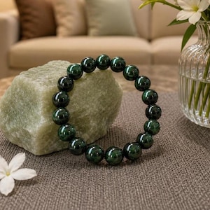 Puede incluir: Una pulsera de cuentas verde oscuro descansa sobre una piedra verde claro. La pulsera está hecha de cuentas grandes y redondas con un acabado brillante. El fondo incluye un jarrón con flores blancas y un sofá de tonos neutros.