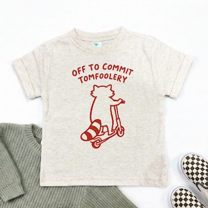 May include: A light gray t-shirt with the text "OFF TO COMMIT TOMFOOLERY" in red. A red raccoon graphic is riding a scooter. A green sweater and black and white checkered shoes are in the background.