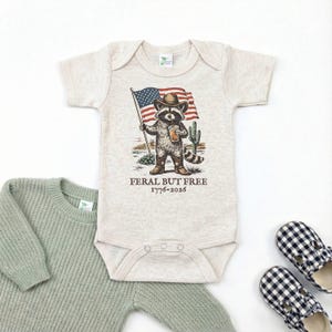 A baby onesie with a raccoon wearing a cowboy hat and holding an American flag, along with a pair of plaid shoes, all laid out on a white background.