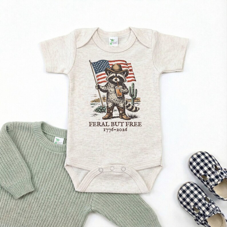 A baby onesie with a raccoon wearing a cowboy hat and holding an American flag, along with a pair of plaid shoes, all laid out on a white background.
