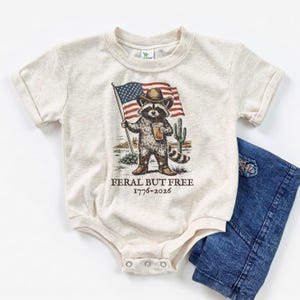 A beige baby onesie with a graphic of a raccoon holding an American flag and the text "Feral But Free 1776-2026" printed on it.