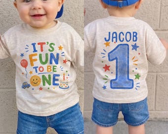 May include: A beige long-sleeve shirt with a blue baseball cap and denim shorts. The front of the shirt features the text IT'S FUN TO BE ONE with colorful graphics of a balloon, smiley face, car, and cake. The back displays the name JACOB and the number 1.