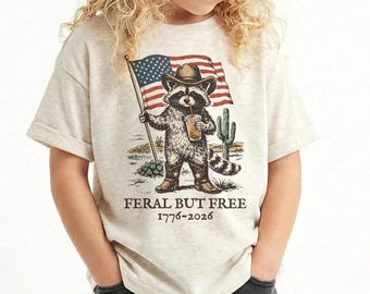 Puede incluir: Camiseta beige con un mapache de dibujos animados con sombrero y botas de vaquero, sosteniendo una bandera americana y una bebida. El texto "FERAL BUT FREE 1776-2026" está impreso debajo. El mapache está en un paisaje desértico.