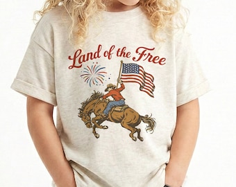 May include: A light beige t-shirt featuring a graphic of a cowboy riding a horse and waving an American flag. The text "Land of the Free" is arched above the graphic in red script. Fireworks are also included in the graphic.