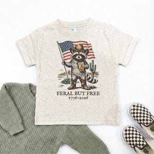 a t-shirt with a graphic of a raccoon holding an American flag and a beer, accompanied by the text "Feral But Free" and a date "1776-2026".