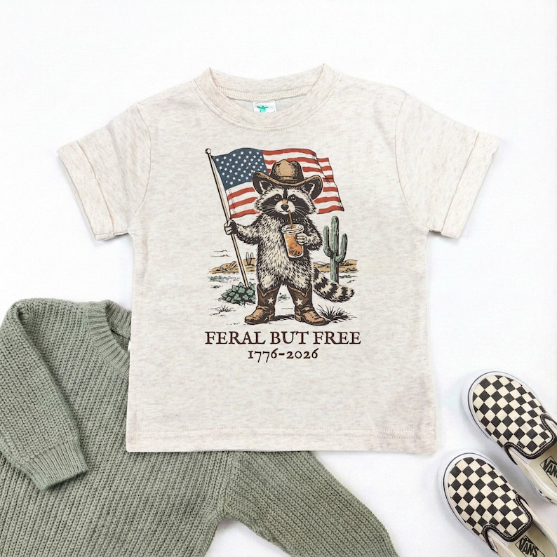 a t-shirt with a graphic of a raccoon holding an American flag and a beer, accompanied by the text "Feral But Free" and a date "1776-2026".