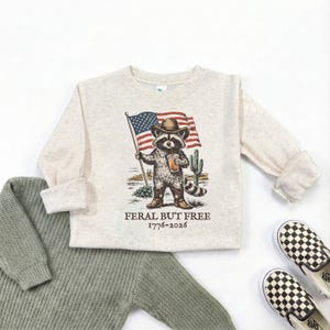 a beige long-sleeved shirt with a graphic of a raccoon holding an American flag, along with a caption that reads "Feral but free 1776-2026".