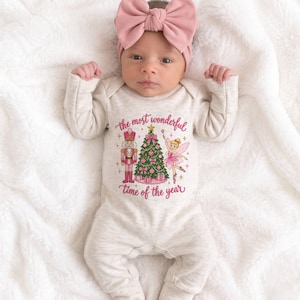 May include: A long-sleeved cream-colored baby bodysuit featuring a graphic of a nutcracker, a decorated Christmas tree, and a fairy with the text "the most wonderful time of the year" in pink, paired with a matching dusty pink fabric bow headband.