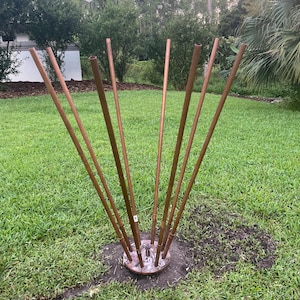 Puede incluir: Una escultura de metal de cobre para exteriores con múltiples tubos verticales que irradian desde una base circular central. La escultura está ubicada en un jardín cubierto de hierba, con árboles y un cielo nublado al fondo.