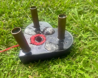 Puede incluir: Un objeto de resina negra en forma de corazón con tres tubos de cobre y un centro enrollado rojo. El objeto tiene piedras incrustadas y un cable rojo que se extiende desde él. El objeto está sobre hierba verde.