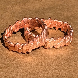 May include: Two copper-colored rings with a textured, braided design. The rings are displayed on a brown surface, showcasing their intricate, twisted pattern. The rings appear to be made of metal and have a warm, reddish-brown hue.