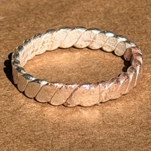 May include: A silver-colored ring with a textured, twisted rope design. The ring is circular and sits on a brown surface. The ring's design features a series of connected, rounded shapes, creating a continuous pattern around the band.