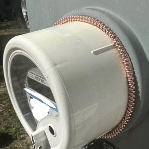 May include: A close-up of a white, cylindrical object with a clear, circular window. A copper coil wraps around the edge of the cylinder. The object is mounted on a light gray surface.