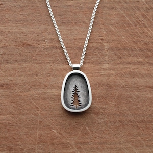 May include: Silver pendant necklace featuring a stylized tree design within an oval frame. The pendant hangs from a delicate silver chain. The necklace is displayed on a wooden surface.