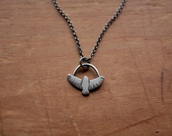 May include: A silver-toned pendant necklace featuring a stylized bird with outstretched wings. The bird is suspended within a circular frame, attached to a delicate chain. The necklace is displayed against a warm, wooden background.