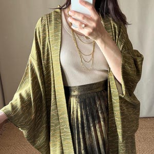 May include: An olive green kimono-style jacket with a flowing, wave-like pattern in a lighter gold color. The jacket is worn over a cream-colored ribbed top and a gold pleated skirt. A layered gold necklace is visible.
