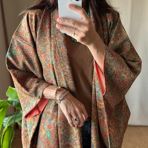 May include: A kimono-style jacket featuring a dense, intricate floral and geometric pattern in shades of rust, gold, teal, and green. The garment has wide sleeves with a solid coral-pink lining and is worn over a plain brown top.