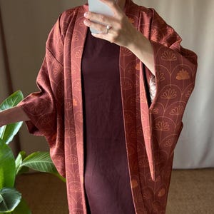 Könnte beinhalten: Ein rostfarbener Kimono mit offener Vorderseite und einem sich wiederholenden Muster aus dünnen, hellen Bogenlinien und stilisierten Blumenmotiven. Das Kleidungsstück hat weite Ärmel und wird über einem dunkelroten ärmellosen Kleid getragen.