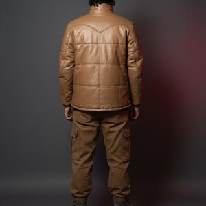 K&ouml;nnte beinhalten: Eine Person tr&auml;gt eine hellbraune Lederjacke und eine passende Cargohose. Die Jacke hat ein gestepptes Design und einen Stehkragen. Die Hose hat mehrere Taschen. Die Person tr&auml;gt auch braune Stiefel.