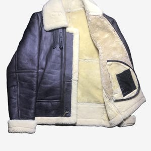 May include: A dark blue leather jacket with a cream-colored shearling lining and collar. The jacket features a zip-up front, two front pockets, and a classic aviator style. The cuffs and hem are trimmed with cream-colored shearling.
