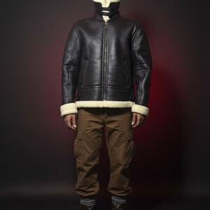 May include: A black leather jacket with a cream-colored shearling lining and trim. The jacket has a zip-up front and two pockets. The person is wearing brown cargo pants and brown boots. The background is a dark red.